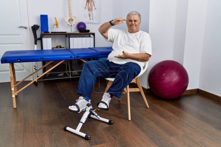 Photo for Senior caucasian man at physiotherapy clinic using pedal exerciser gesturing with hands showing big and large size sign, measure symbol. smiling looking at the camera. measuring concept. - Royalty Free Image