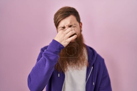 Téléchargez les photos : Caucasian man with long beard standing over pink background smelling something stinky and disgusting, intolerable smell, holding breath with fingers on nose. bad smell - en image libre de droit