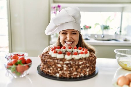 Foto de Joven hermosa mujer hispana sonriendo confiada oliendo pastel en la cocina - Imagen libre de derechos