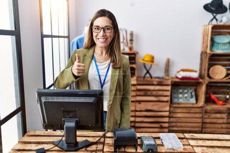Photo for Young hispanic woman working as manager at retail boutique smiling happy and positive, thumb up doing excellent and approval sign - Royalty Free Image