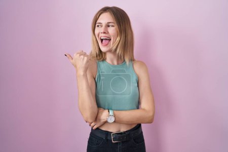 Foto de Blonde caucasian woman standing over pink background smiling with happy face looking and pointing to the side with thumb up. - Imagen libre de derechos