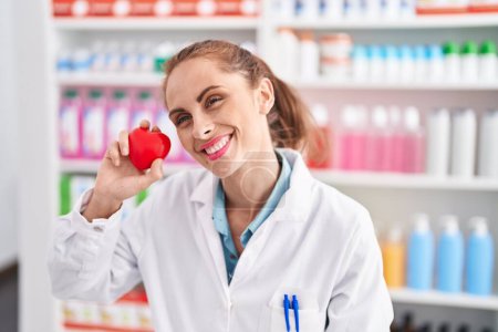 Téléchargez les photos : Young beautiful hispanic woman pharmacist smiling confident holding heart at pharmacy - en image libre de droit