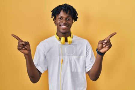 Téléchargez les photos : Jeune homme africain avec dreadlocks debout sur fond jaune souriant confiant pointant avec les doigts vers différentes directions. espace de copie pour la publicité - en image libre de droit
