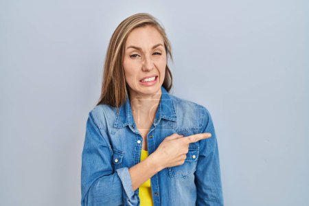 Foto de Young blonde woman standing over blue background pointing aside worried and nervous with forefinger, concerned and surprised expression - Imagen libre de derechos