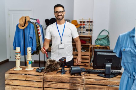 Foto de Joven comerciante hispano sonriendo confiado limpiando polvo en la tienda de ropa - Imagen libre de derechos
