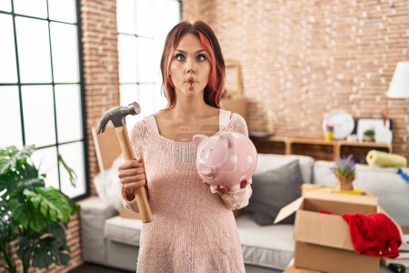Téléchargez les photos : Young caucasian woman holding piggy bank at new home making fish face with mouth and squinting eyes, crazy and comical. - en image libre de droit