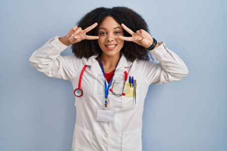 Photo for Young african american woman wearing doctor uniform and stethoscope doing peace symbol with fingers over face, smiling cheerful showing victory - Royalty Free Image
