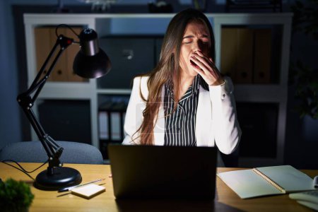 Téléchargez les photos : Jeune femme brune travaillant au bureau la nuit avec un ordinateur portable ennuyé bâillant fatigué couvrant la bouche avec la main. agitation et somnolence. - en image libre de droit
