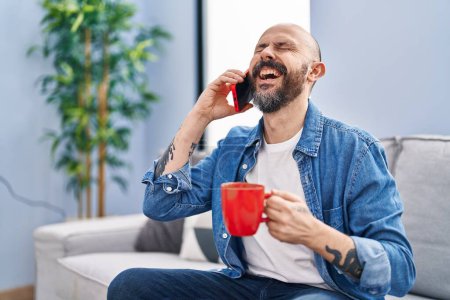Foto de Joven hombre calvo hablando en el teléfono inteligente beber café en casa - Imagen libre de derechos