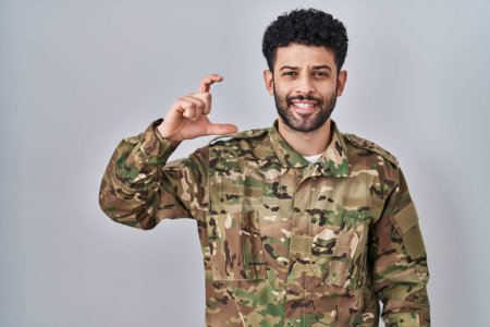 Téléchargez les photos : Homme arabe portant uniforme de camouflage armée geste souriant et confiant avec la main faisant signe de petite taille avec les doigts à la recherche et la caméra. concept de mesure. - en image libre de droit