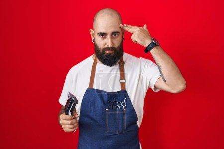 Foto de Young hispanic man with beard and tattoos wearing barber apron holding razor shooting and killing oneself pointing hand and fingers to head like gun, suicide gesture. - Imagen libre de derechos