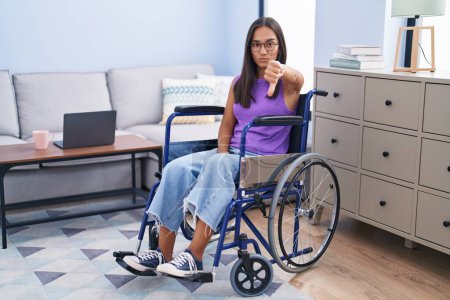 Foto de Young hispanic woman sitting on wheelchair at home looking unhappy and angry showing rejection and negative with thumbs down gesture. bad expression. - Imagen libre de derechos