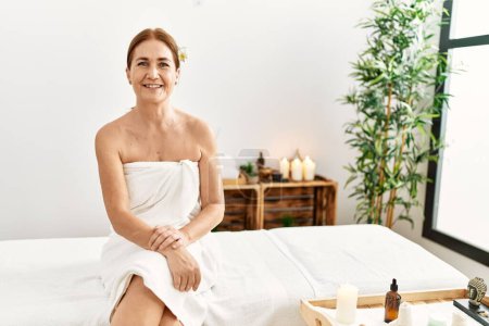Téléchargez les photos : Moyen âge femme caucasienne portant serviette assis sur le conseil de massage au centre de beauté - en image libre de droit
