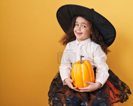 Foto de Adorable hispanic girl wearing halloween costume holding pumpkin over isolated yellow background - Imagen libre de derechos