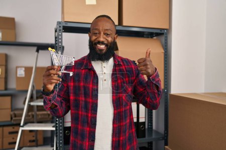 Foto de African american man working at small business ecommerce holding cart smiling happy and positive, thumb up doing excellent and approval sign - Imagen libre de derechos