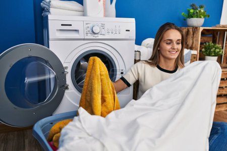 Foto de Joven mujer rubia sonriendo confiada sosteniendo ropa limpia en la lavandería - Imagen libre de derechos