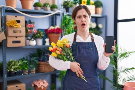 Téléchargez les photos : Femme brune travaillant dans un magasin de fleuristes tenant son smartphone sous le choc, sceptique et sarcastique, surprise avec la bouche ouverte - en image libre de droit