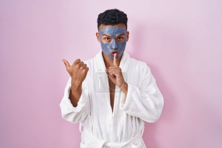 Photo for Young hispanic man wearing beauty face mask and bath robe asking to be quiet with finger on lips pointing with hand to the side. silence and secret concept. - Royalty Free Image