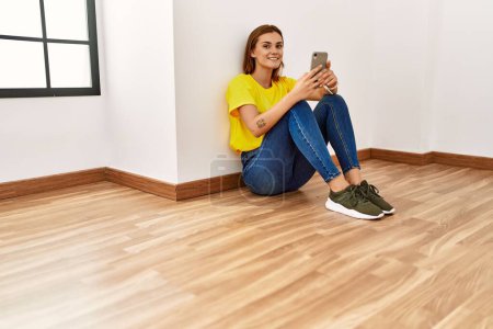 Téléchargez les photos : Jeune femme utilisant smartphone assis sur le sol dans une pièce vide - en image libre de droit