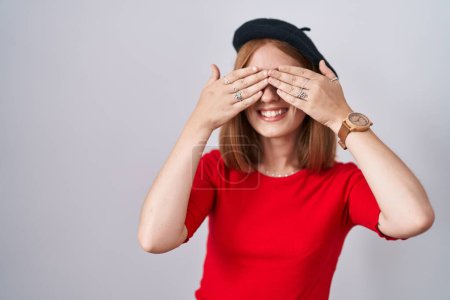 Téléchargez les photos : Jeune femme rousse debout portant des lunettes et béret couvrant les yeux avec les mains souriantes gaies et drôles. concept aveugle. - en image libre de droit