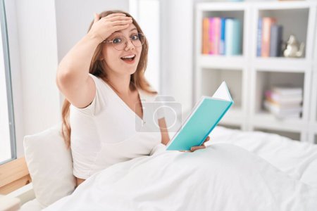 Téléchargez les photos : Jeune femme caucasienne assise sur le lit à la maison lisant un livre stressé et frustré avec la main sur la tête, visage surpris et en colère - en image libre de droit