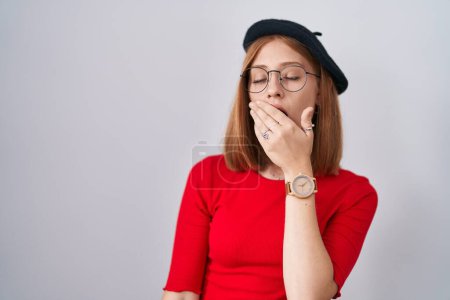 Foto de Mujer pelirroja joven de pie con gafas y boina aburrida bostezando cansada cubriendo la boca con la mano. inquietud y somnolencia. - Imagen libre de derechos