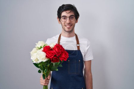 Foto de Young hispanic man holding bouquet of white and red roses with a happy and cool smile on face. lucky person. - Imagen libre de derechos