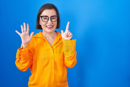 Foto de Mujer hispana de mediana edad con gafas de pie sobre fondo azul mostrando y apuntando hacia arriba con los dedos número siete mientras sonríe confiada y feliz. - Imagen libre de derechos