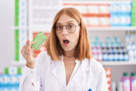 Foto de Young redhead woman working at pharmacy drugstore holding birth control pills scared and amazed with open mouth for surprise, disbelief face - Imagen libre de derechos