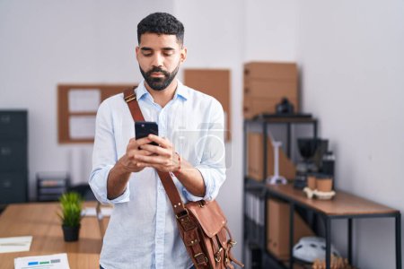 Photo for Hispanic man with beard using smartphone at the office relaxed with serious expression on face. simple and natural looking at the camera. - Royalty Free Image