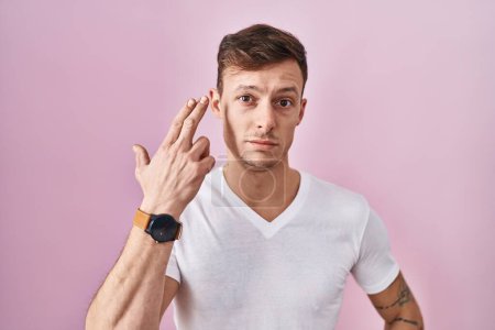 Foto de Hombre caucásico de pie sobre fondo rosa disparando y matándose señalando la mano y los dedos a la cabeza como arma, gesto suicida. - Imagen libre de derechos
