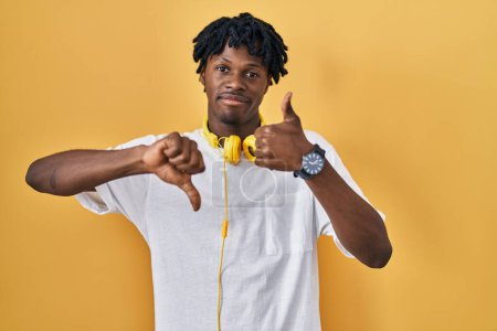 Téléchargez les photos : Jeune homme africain avec dreadlocks debout sur fond jaune faisant pouces vers le haut et vers le bas, désaccord et expression d'accord. conflit fou - en image libre de droit