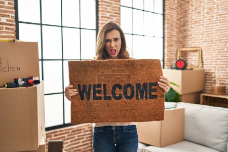 Foto de Young woman holding welcome doormat at new home clueless and confused expression. doubt concept. - Imagen libre de derechos