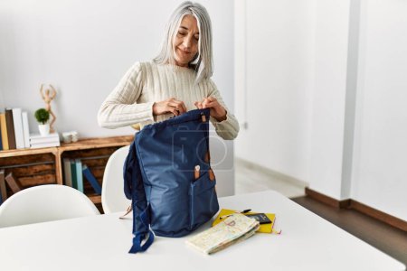 Foto de Mujer de pelo gris de mediana edad sonriendo confiada preparando la mochila de viaje en casa - Imagen libre de derechos