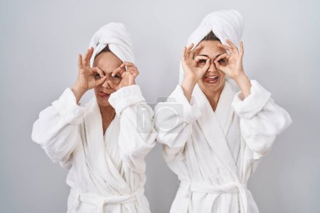 Foto de Mujer e hija de mediana edad con albornoz blanco y toalla haciendo buen gesto como prismáticos sacando la lengua, ojos mirando a través de los dedos. expresión loca. - Imagen libre de derechos