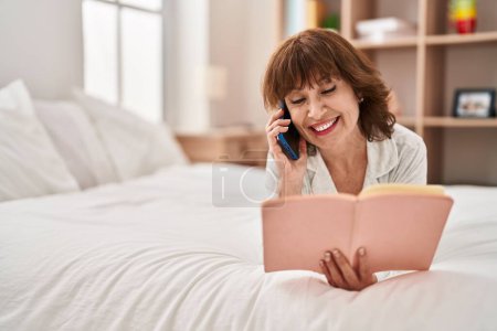 Photo for Middle age woman talking on smartphone reading book at bedroom - Royalty Free Image