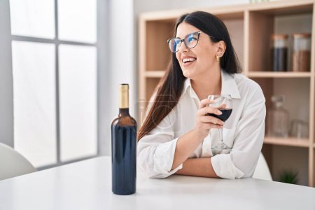 Foto de Mujer hispana joven bebiendo copa de vino sentada en la mesa en casa - Imagen libre de derechos