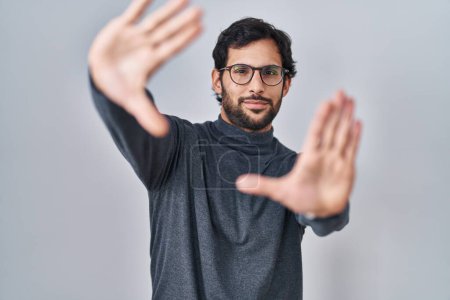 Foto de Handsome latin man standing over isolated background doing frame using hands palms and fingers, camera perspective - Imagen libre de derechos