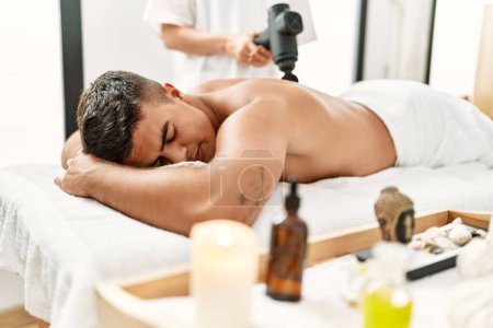 Photo for Young hispanic man relaxed having back massage using percussion pistol at beauty center - Royalty Free Image