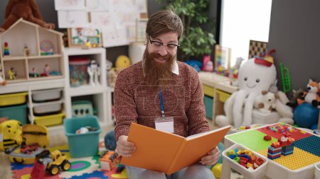 Photo for Young redhead man preschool teacher reading book sitting on chair at kindergarten - Royalty Free Image