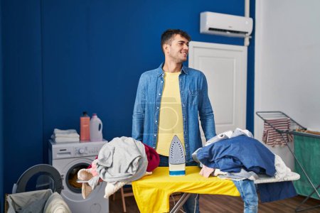 Foto de Hombre hispano joven planchando ropa en casa mirando al costado, pose de perfil relajado con rostro natural y sonrisa confiada. - Imagen libre de derechos