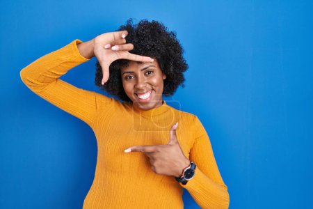 Foto de Black woman with curly hair standing over blue background smiling making frame with hands and fingers with happy face. creativity and photography concept. - Imagen libre de derechos