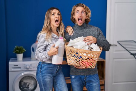 Photo for Couple holding laundry basket and detergent bottle angry and mad screaming frustrated and furious, shouting with anger looking up. - Royalty Free Image