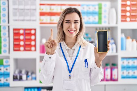 Photo for Young beautiful woman working at pharmacy drugstore showing smartphone screen smiling with an idea or question pointing finger with happy face, number one - Royalty Free Image