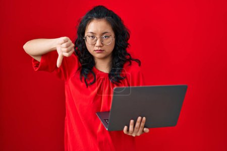 Téléchargez les photos : Jeune femme asiatique travaillant à l'aide d'un ordinateur portable avec le visage en colère, signe négatif montrant aversion avec les pouces vers le bas, concept de rejet - en image libre de droit