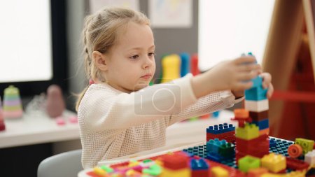 Téléchargez les photos : Adorable blonde girl playing with construction blocks sitting on table at kindergarten - en image libre de droit