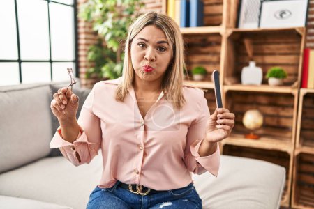 Téléchargez les photos : Young hispanic woman holding eye lashes curler and nail file puffing cheeks with funny face. mouth inflated with air, catching air. - en image libre de droit