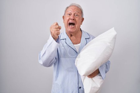 Foto de Senior man with grey hair wearing pijama hugging pillow angry and mad raising fist frustrated and furious while shouting with anger. rage and aggressive concept. - Imagen libre de derechos