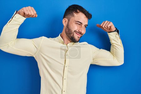 Téléchargez les photos : Handsome hispanic man standing over blue background showing arms muscles smiling proud. fitness concept. - en image libre de droit