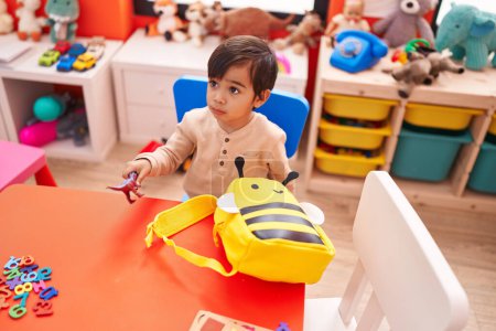 Foto de Adorable estudiante hispano sentado en la mesa jugando con un juguete de dino en el jardín de infantes - Imagen libre de derechos